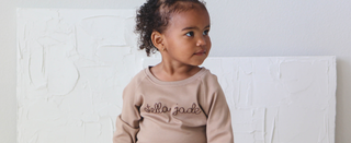 A toddler girl sits on a stool while wearing a hand-embroidered mauve pullover with her name "Stella Jade" on the front in cursive.