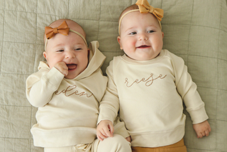 Two infants wear cream colored hand-embroidered outfits. One is a hoodie with the name "Eden" in cursive and one is a pullover with the name "reese" in cursive.