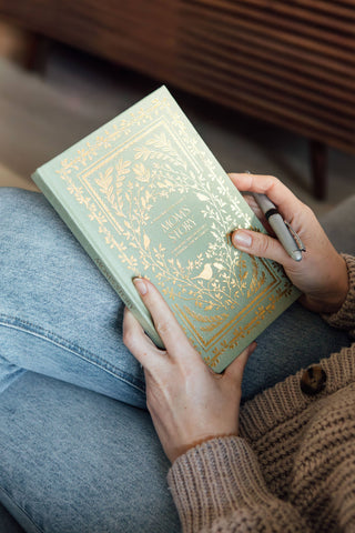 Person holding a green book with gold detailing and a pen, sitting on a wooden bench.