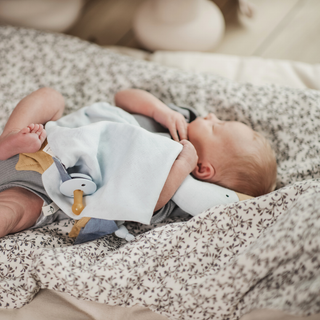 A baby lying on a patterned changing pad or blanket, wrapped in a white cloth with a pacifier nearby.