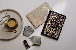 Decorative books with ornate covers, a cup of coffee, and glasses on a light surface.