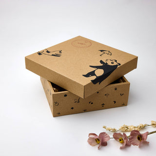 A brown kraft paper gift box with a matching lid, decorated with black cartoon animal illustrations and a small round logo stamp. The box is shown slightly open with dried pink flowers placed in front on a clean white background.