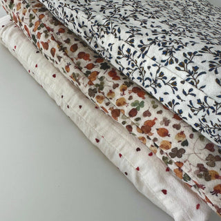 Three folded quilted blankets with different floral and botanical patterns stacked diagonally on a light gray surface.