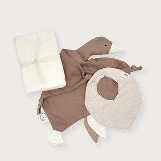 A baby comforter set featuring a duck-shaped lovey in taupe and cream, a matching folded white blanket tied with a pale pink ribbon, and a light beige bib. The items are arranged on a solid light gray background.