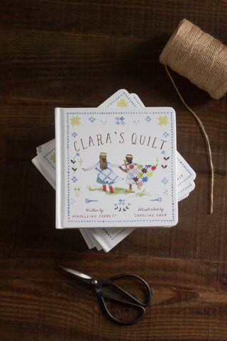Stack of 'Clara's Quilt' books on a wooden surface with scissors and twine.