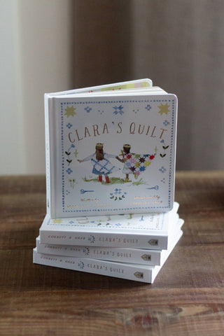 Stack of books titled 'Clara's Quilt' on a wooden surface with a neutral background
