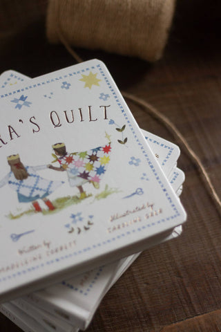 A stack of square quilted fabric boxes with illustrated covers reading 'Aria's Quilt,' photographed on a dark wooden surface with a burlap roll blurred in the background.