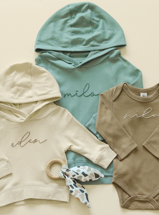 Two hand-embroidered hoodies for children and a long-sleeve hand-embroidered body suit. The hoodies are teal and cream colored while the bodysuit is a light brown.