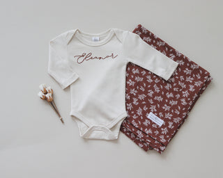Long sleeve infant bodysuit with hand-embroidered name in the center. Also shown is a neatly folded burgundy swaddle with a white floral print.