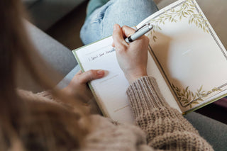 Person writing in a notebook with a pen, wearing a brown sweater.