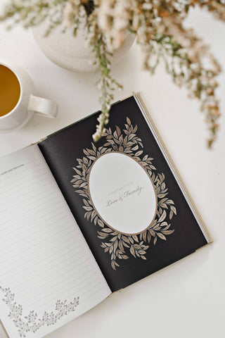 Open book with decorative cover on a white surface with a cup of coffee and flowers.