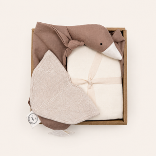 A baby gift set in a brown cardboard box featuring a plush duck toy in taupe and cream, an ivory swaddle wrap, and an off-white knit bib. 