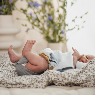 A baby lying on their back on a patterned blanket, wearing a light blue and white outfit with gray diaper cover, in a softly lit room with blurred flowers in the background.